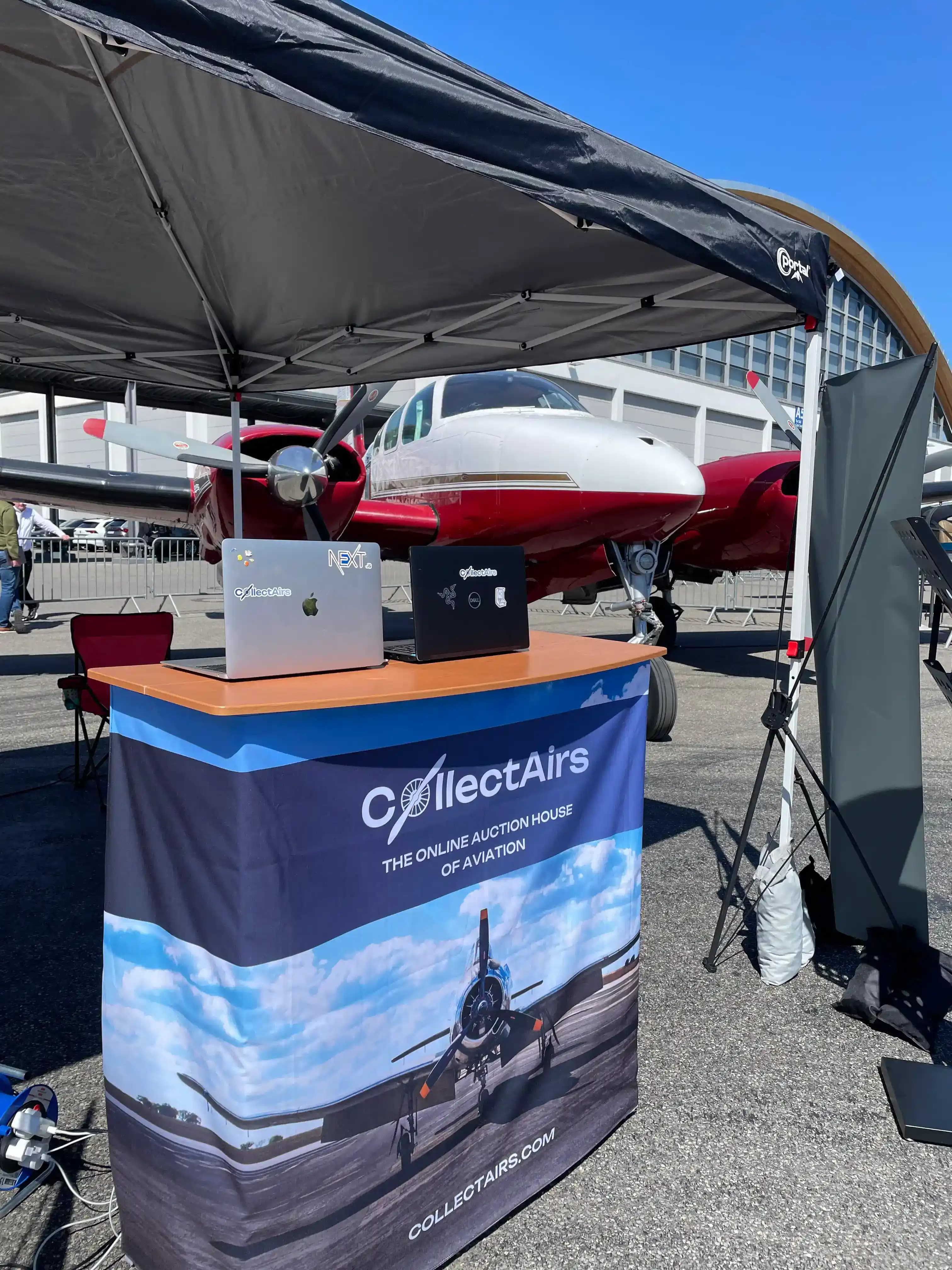 CollectAirs branded counter with laptops at AERO booth in front of twin-engine aircraft