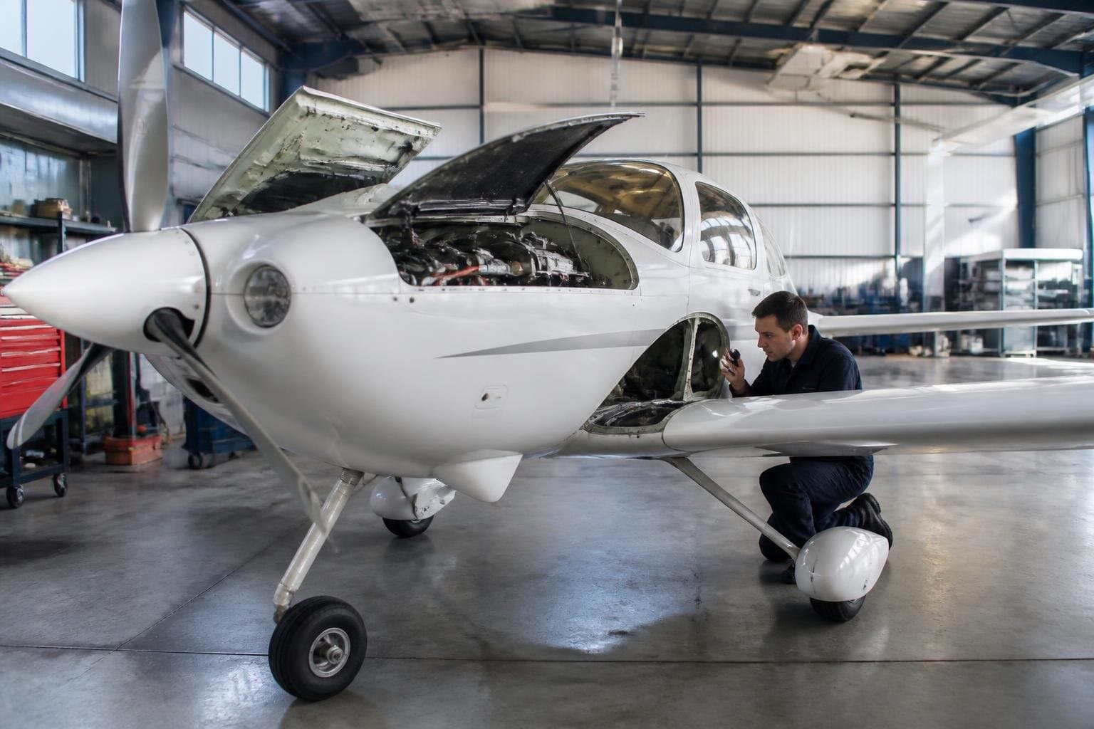 Technician inspecting the wing root of a light aircraft with access panels open in a bright maintenance hangar