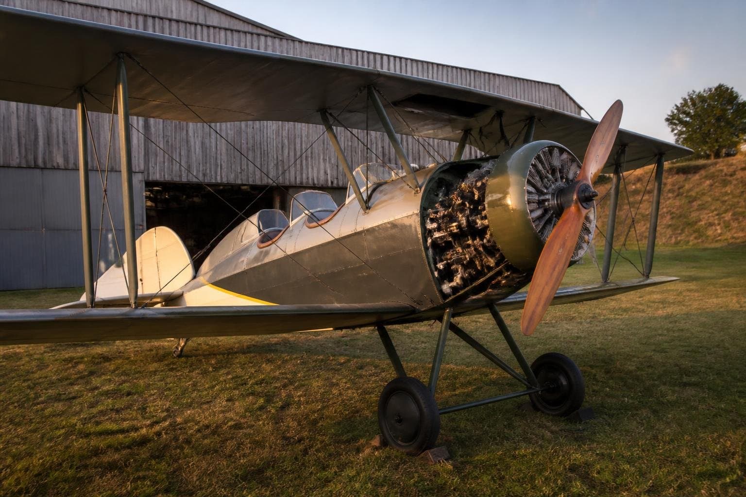 Classic open-cockpit biplane with an exposed seven-cylinder radial engine outside a heritage hangar at golden hour
