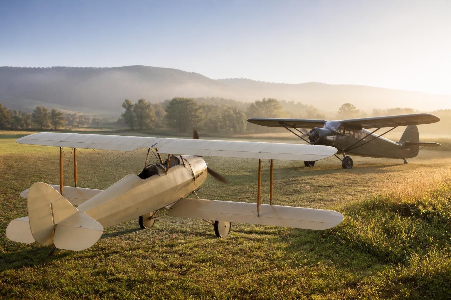 Two classic fabric-covered taildraggers on a sunlit grass airstrip with rolling hills behind